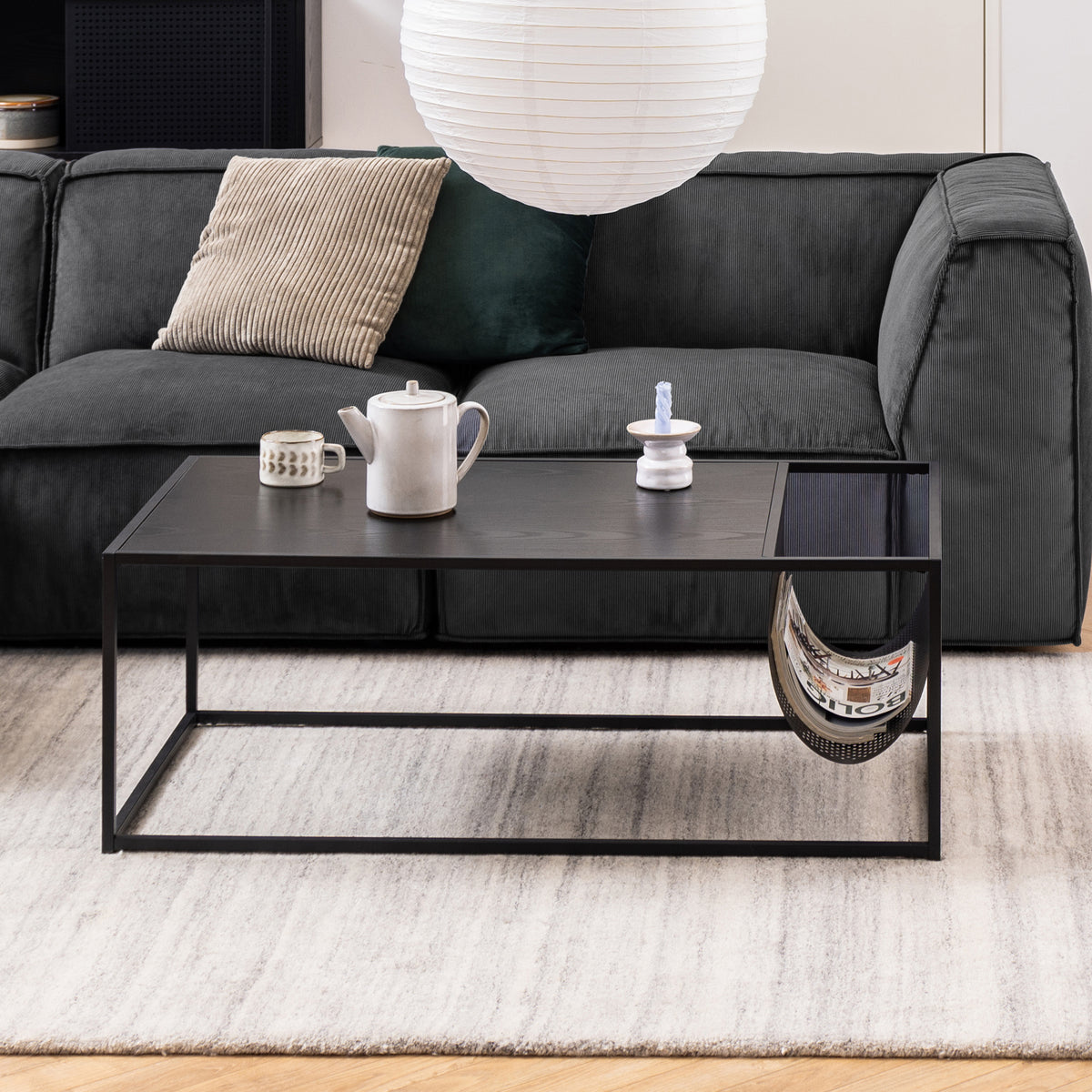 A Skeleto coffee table (110x40) in front of a graphite-coloured sofa on a light-coloured carpet, with a jug and magazines in a metal pocket