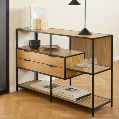 A low Skeleto bookcase (114x78) in Wild Oak under a black lamp, with books, bowls and glasses on the shelves
